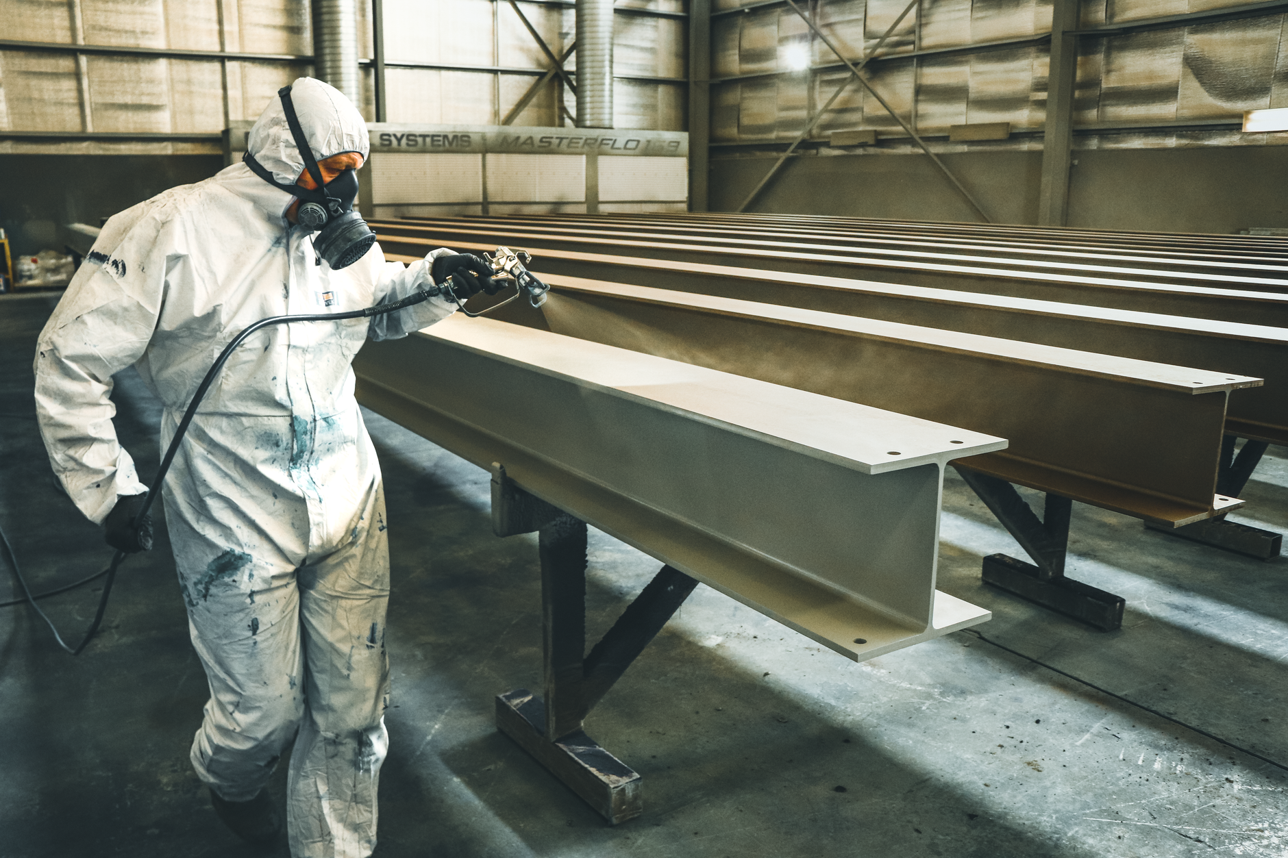 Pracownik w kombinezonie maluje natryskowo metalowe belki w hali
